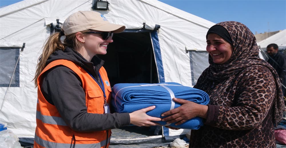 Eye-level view of relief worker handing blue wool blanket to elderly refugee woman at aid distribution point, blue sky, showing compassionate humanitarian relief supply delivery for vulnerable populations