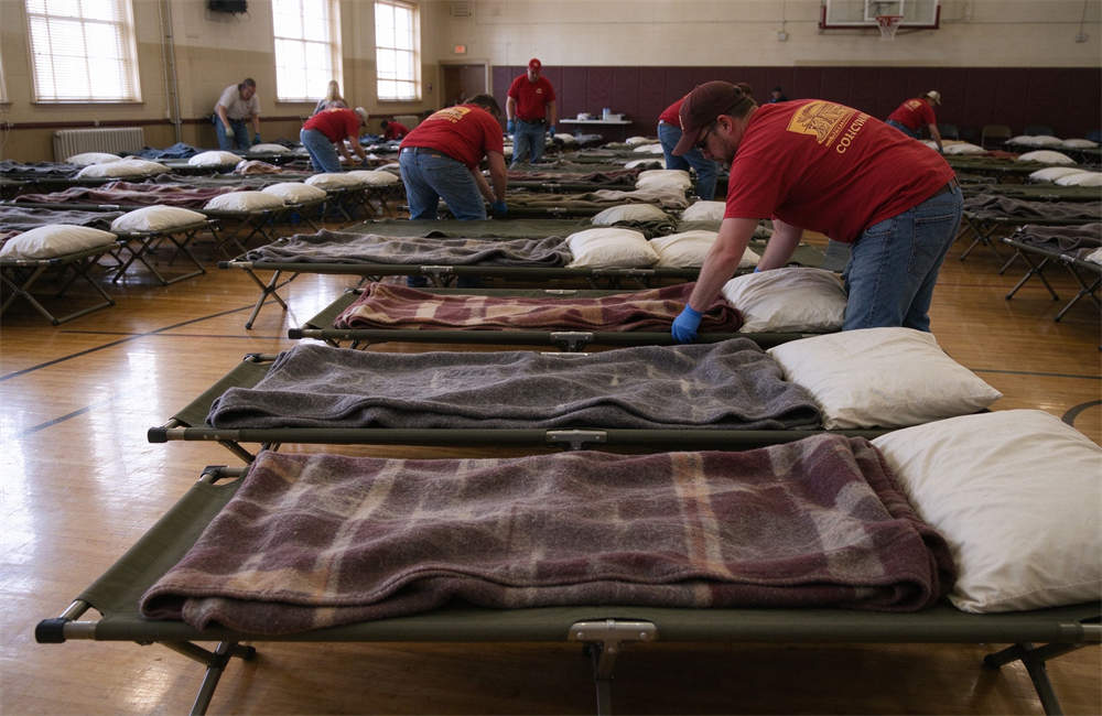 Rows of folding cots in community shelter gymnasium with wool blankets and pillows, emergency shelter bedding supplies for high wind disaster displaced residents