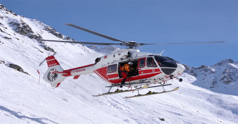 Wide view of red rescue helicopter hovering over snowy mountain during avalanche rescue showing aerial support